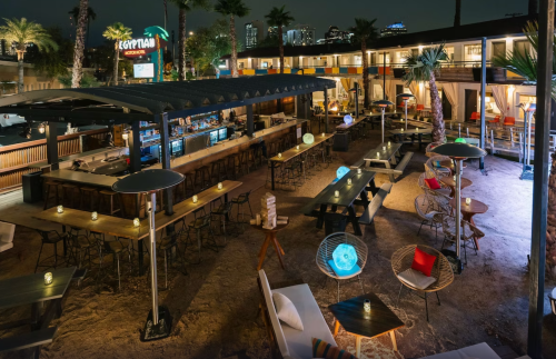 A vibrant outdoor bar area at night, featuring palm trees, seating, and colorful lights in a lively atmosphere.