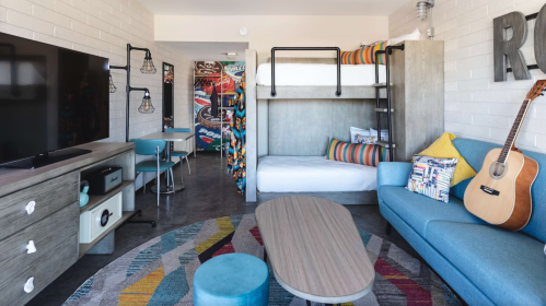A modern hotel room featuring a bunk bed, sofa, TV, and colorful decor with a guitar on the floor.