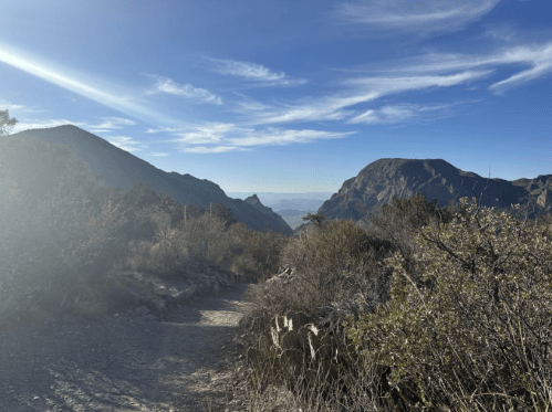 A scenic mountain trail with a clear blue sky and distant peaks under soft sunlight.
