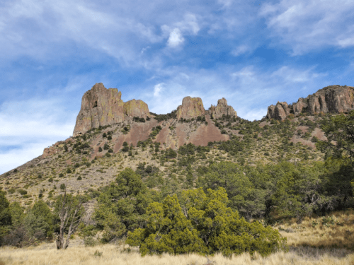 A rugged mountain landscape with rocky peaks and green shrubs under a blue sky with scattered clouds.