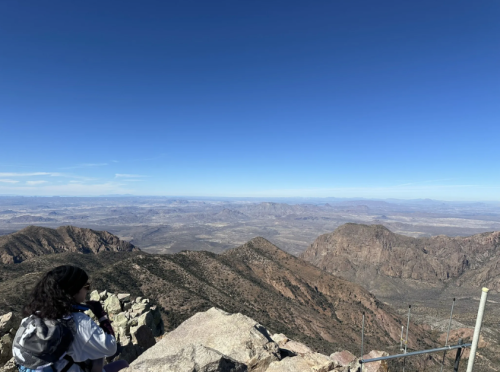 A person stands on a rocky mountain peak, overlooking a vast landscape of mountains and valleys under a clear blue sky.