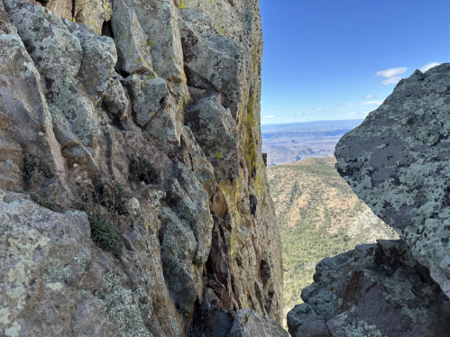 A narrow rocky crevice between steep cliffs, with distant mountains and a clear blue sky in the background.