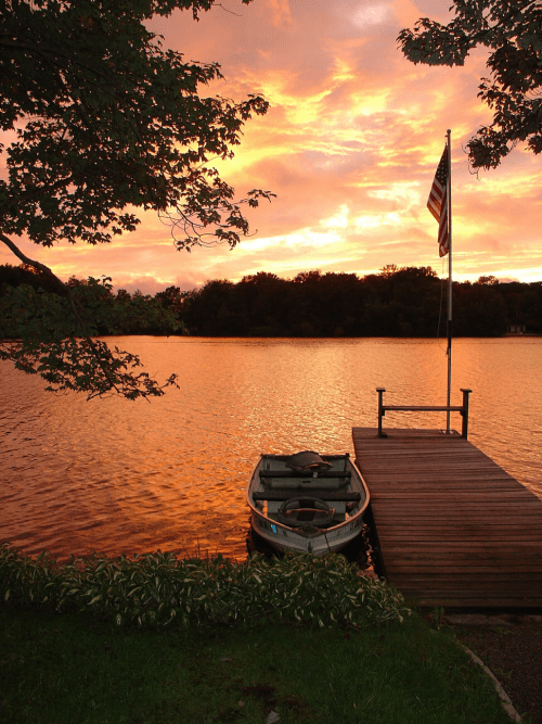 A serene lake at sunset, with vibrant orange skies, a small boat by a wooden dock, and an American flag waving nearby.