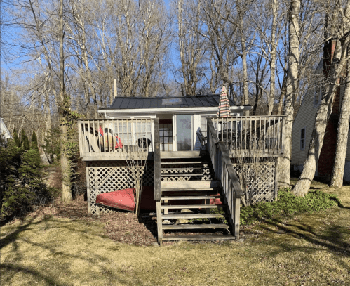A small house with a deck and stairs, surrounded by trees and grass on a sunny day.
