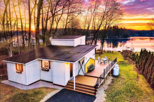 A cozy house by a lake at sunset, surrounded by trees and featuring a deck with outdoor seating.