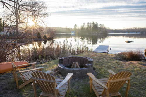 A serene lakeside scene at sunrise, featuring a fire pit and wooden chairs overlooking calm water and lush greenery.