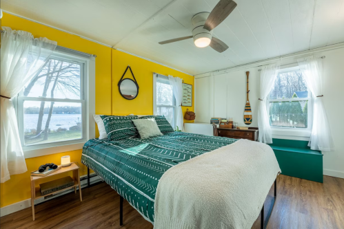 Cozy bedroom with a green and white bedspread, yellow walls, and a view of a lake through large windows.
