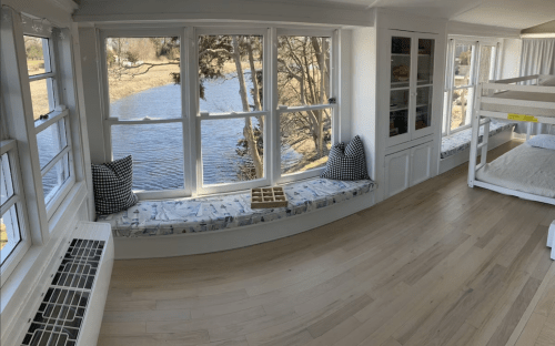 Bright room with large windows overlooking a river, featuring cozy window seats and a bunk bed.