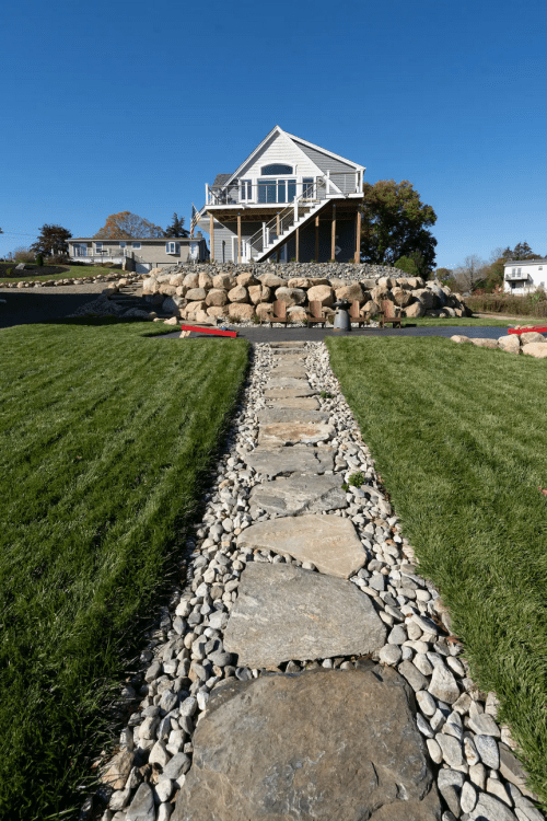 A stone pathway leads to a modern house on a raised platform, surrounded by grass and large rocks under a clear blue sky.