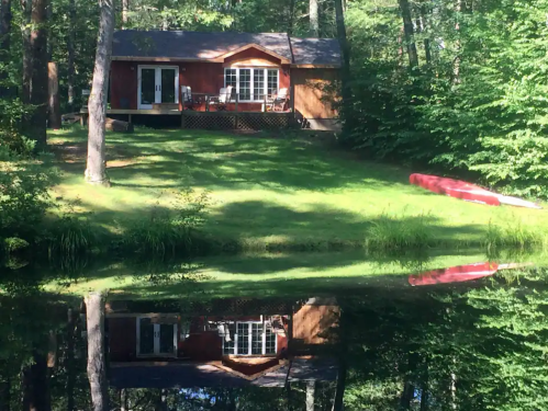A cozy cabin surrounded by trees, reflecting in calm water, with a red canoe nearby on the shore.
