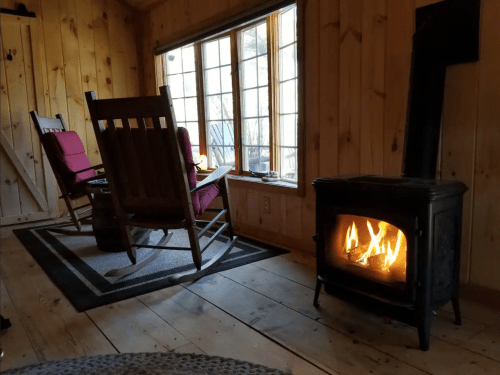 Cozy interior with wooden walls, two rocking chairs, and a glowing stove by a large window.