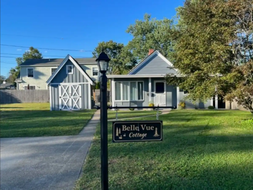 A charming cottage with a sign reading "Belle Vue Cottage," surrounded by green grass and trees under a clear blue sky.
