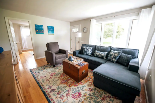 Cozy living room with a blue sectional sofa, armchair, coffee table, and colorful rug, featuring large windows and natural light.