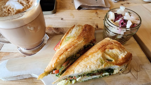 A toasted sandwich with vegetables and a small jar of fruit salad, accompanied by a latte on a wooden table.