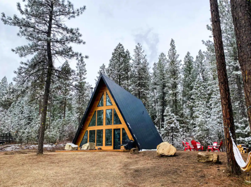 A cozy A-frame cabin surrounded by tall pine trees and a snowy landscape, with red chairs in front.