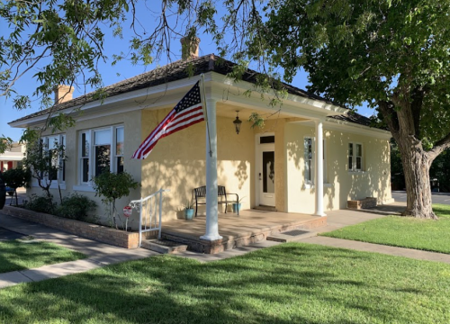 A yellow house with a porch, American flag, and green lawn under a clear blue sky.