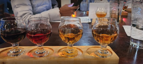 A wooden flight board with four glasses of different colored spirits, set on a table in a dimly lit bar.
