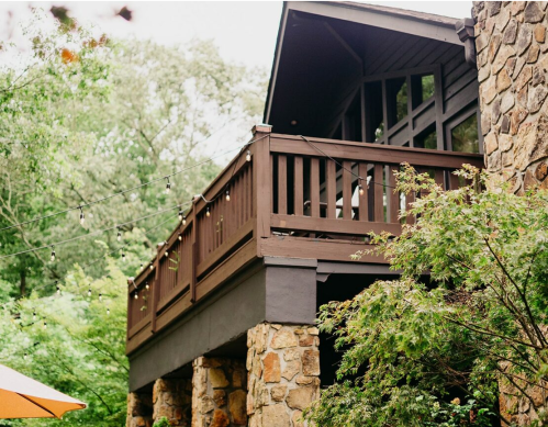 A wooden balcony with string lights, supported by stone pillars, surrounded by greenery.