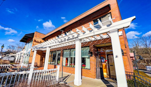 A brick restaurant with a white pergola entrance, surrounded by outdoor seating and a clear blue sky.