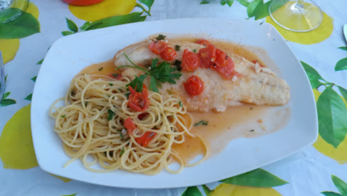 A plate of fish topped with tomatoes and herbs, served alongside a portion of spaghetti on a lemon-patterned tablecloth.