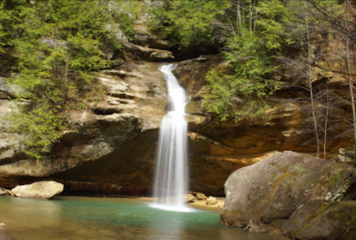 A serene waterfall cascades into a clear pool, surrounded by lush greenery and rocky cliffs.