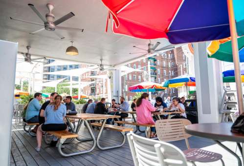 A lively outdoor dining area with colorful umbrellas, featuring groups of people enjoying meals at picnic tables.