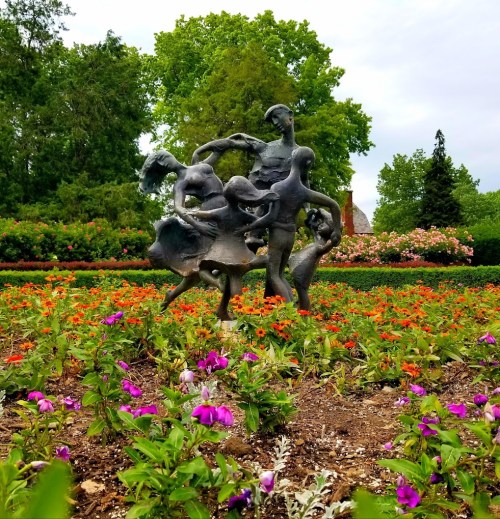 A bronze sculpture of dancing figures surrounded by colorful flowers in a lush garden.
