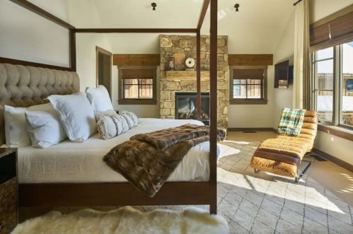 Cozy bedroom featuring a four-poster bed, stone fireplace, and a stylish lounge chair by a large window.