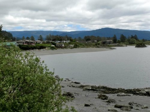 A serene coastal view with houses along the shore, rocky beach, and mountains in the background under a cloudy sky.