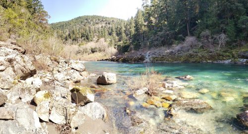 A serene river flows through a rocky landscape, surrounded by lush green trees and a clear blue sky.