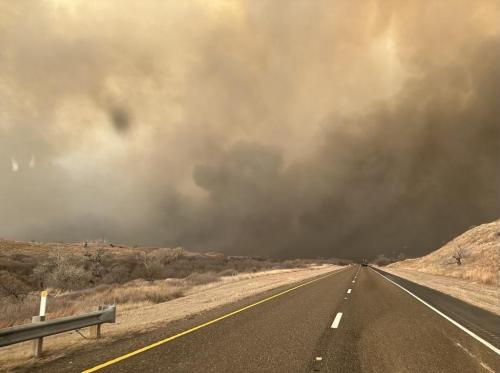 A highway scene with dark smoke billowing in the sky, indicating a nearby fire, surrounded by dry landscape.