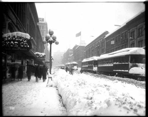 A snowy street scene with vintage streetcars, pedestrians, and buildings, capturing a winter day in the past.