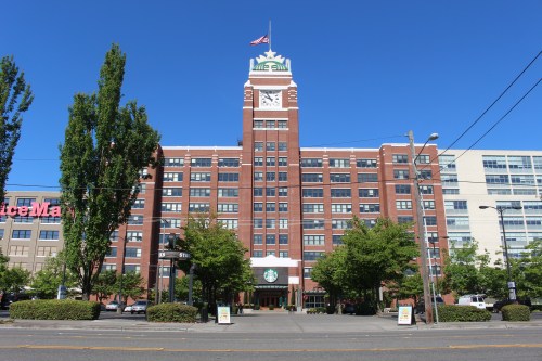 Historic brick building with a clock tower, featuring a Starbucks entrance and surrounded by trees and clear blue sky.