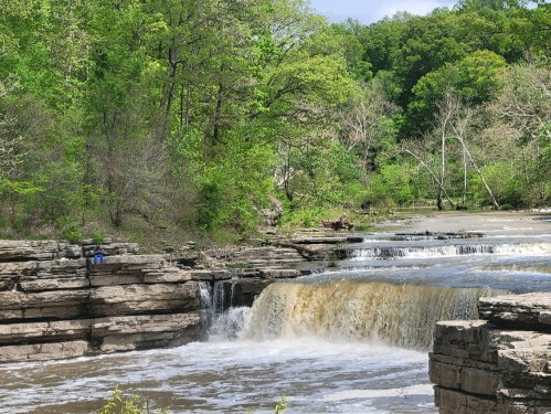 A serene waterfall cascades over rocky ledges, surrounded by lush green trees and a peaceful natural landscape.