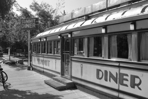 A vintage diner with a curved roof and large windows, featuring a sign that reads "Summit Diner" in a tree-lined area.