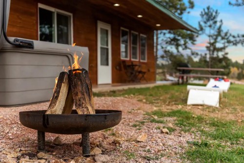 A small fire burns between two logs in a metal fire pit, with a cabin and picnic area in the background.