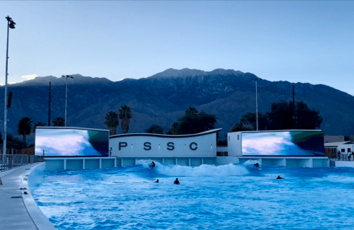 A wave pool with large screens, surrounded by mountains and palm trees, under a clear blue sky.
