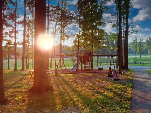 A playground with slides and swings, surrounded by trees, illuminated by the sun setting in the background.