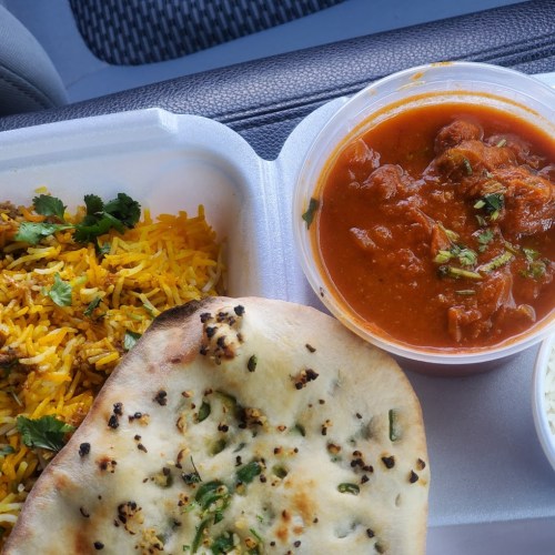 A takeout container with biryani rice, naan bread, and a bowl of curry topped with herbs.
