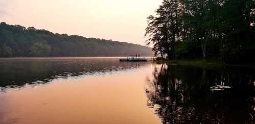 A serene lake at sunrise, with a dock and trees reflecting on the calm water.