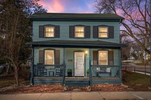 A two-story blue house with white trim, front porch, and windows lit up at dusk, surrounded by trees and a sidewalk.