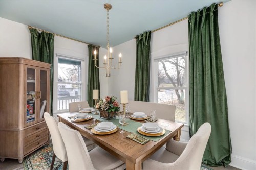 A cozy dining room with a wooden table set for four, green curtains, and a chandelier, overlooking a scenic view.