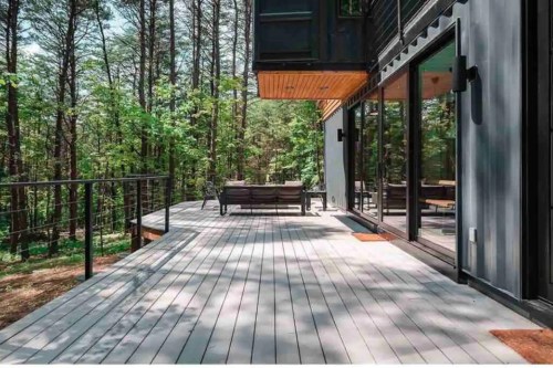 A modern deck surrounded by trees, featuring a seating area and large glass windows reflecting the natural scenery.