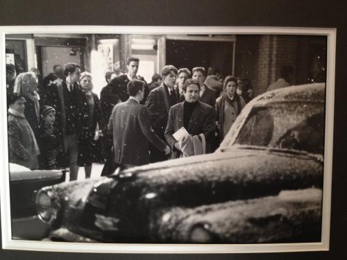 A black-and-white photo of a snowy street scene with people walking and a vintage car parked nearby.