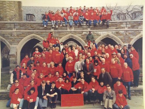 A large group of people in red sweatshirts poses together outside a stone building, smiling for the camera.