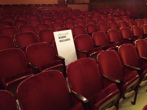 A row of empty red theater seats with a sign that reads "Reserved for Robin Williams" prominently displayed.