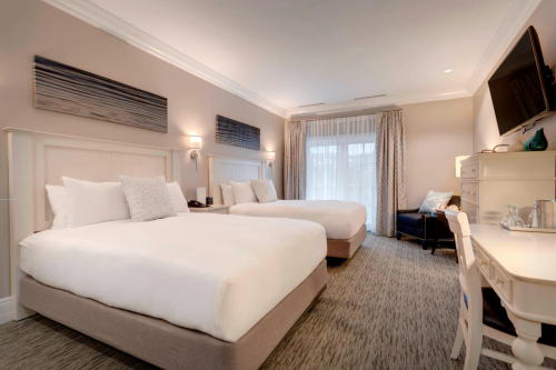 A spacious hotel room featuring two large beds, a desk, and a seating area with natural light from a window.