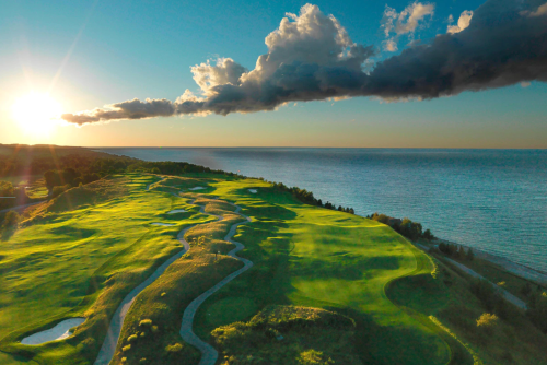 Aerial view of a lush golf course by the ocean at sunset, with dramatic clouds and vibrant green fairways.