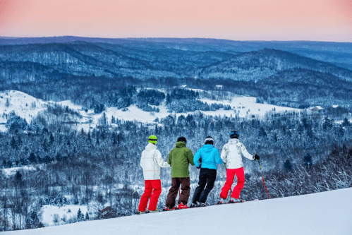 Four skiers in colorful gear stand on a snowy slope, overlooking a vast winter landscape at sunset.
