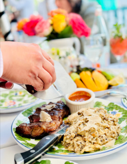 A hand drizzles sauce over a steak and risotto, with colorful flowers and drinks in the background.